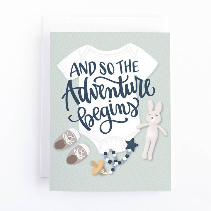 Greeting card featuring an illustration of a babygrow, baby shoes, a pacifier and a stuffed toy on a light green coloured background with a dotted pattern. The text 'And so the adventure begins' is printed over the image. 