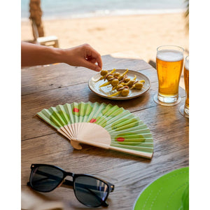 Image showing an open fan with a green and red olive design lying on a table in a beach scene. It is placed beside a plate of olives, some beer and a pair of sunglasses.