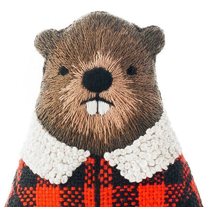 A close-up of an embroidered beaver plush toy with brown stitching for fur, a black nose, white teeth, a white textured collar, and a red-and-black plaid body, shown against a plain white background.