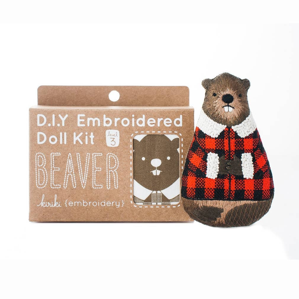 A DIY embroidered doll kit box next to a finished beaver doll wearing a red and black plaid sweater with a white collar, on a white background. The box is brown with white text. 