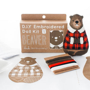 A DIY embroidered doll kit box next to a finished beaver doll wearing a red and black plaid sweater with a white collar, on a white background. The box is brown with white text. An embroidery pattern and some thread sit in front of the box and beaver. 