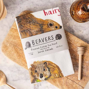 Image of a folded white cotton tea towel showing beaver illustrations with text printed underneath. a paper labe with the heading 'beavers' is wrapped around the middle of the towel. It lies on a wooden board. 