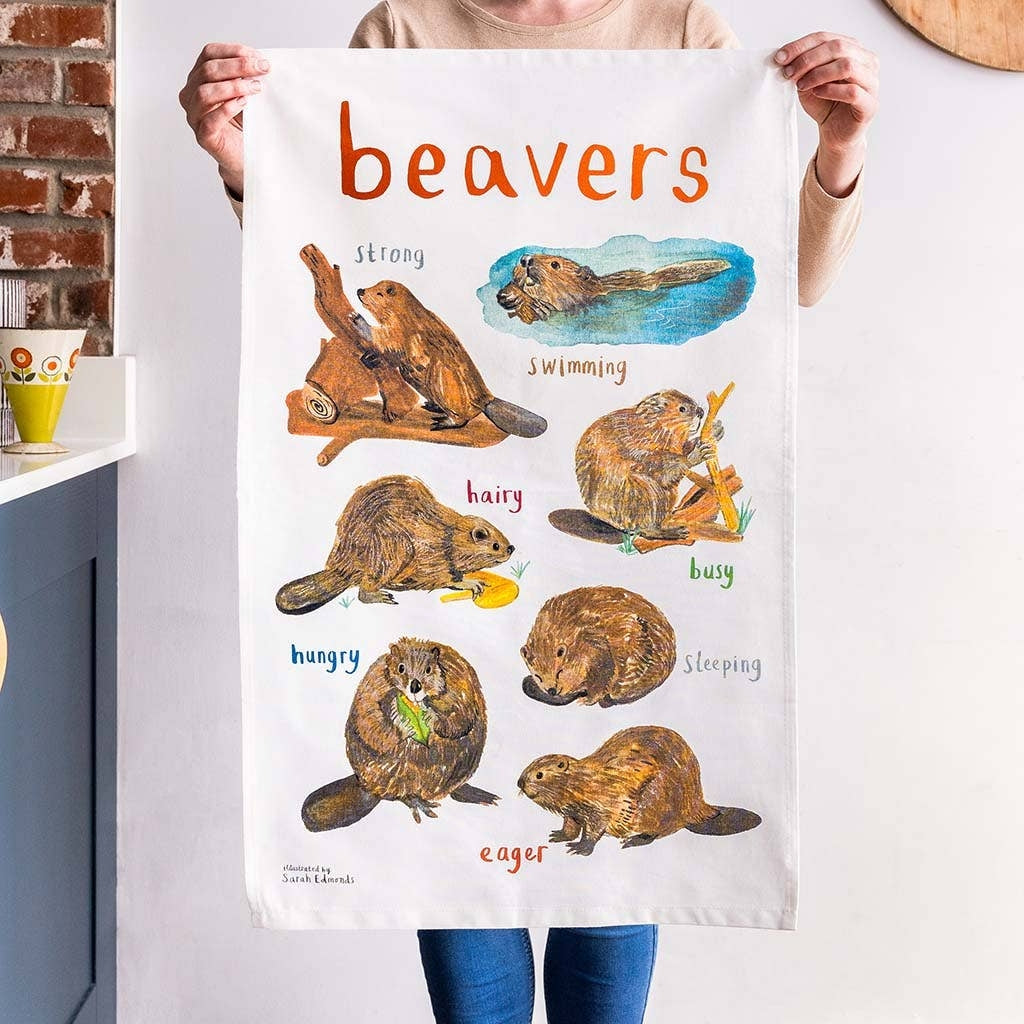 A person is holding up a white cotton tea towel showing beaver illustrations with text printed underneath. The heading 'beavers' is printed at the top. 