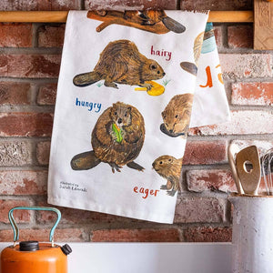 Image of a folded white cotton tea towel showing beaver illustrations with text printed underneath. The towel hangs on a wooden dowel against a red brick background. 