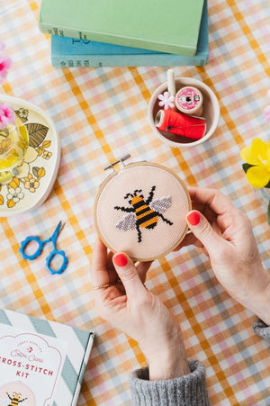Person holding a cross-stitched bee contained in a circular embroidery hoop on a checkered tablecloth with books and craft items.