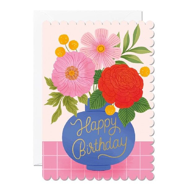 Greeting card with a scalloped edge featuring an illustration of a blue vase holding a bunch of flowers on a pink checkered surface against a light pink background. The text 'Happy Birthday' is printed on the vase. 