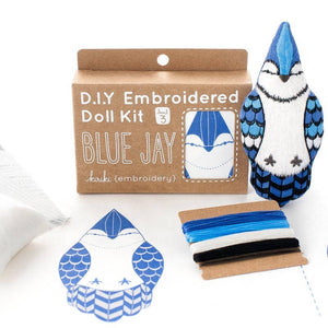 A DIY embroidered doll kit box next to a finished blue jay plush doll on a white background. The box is brown with white text. An embroidery pattern for the blue jay and some threads sit in front of the box and finished product. 