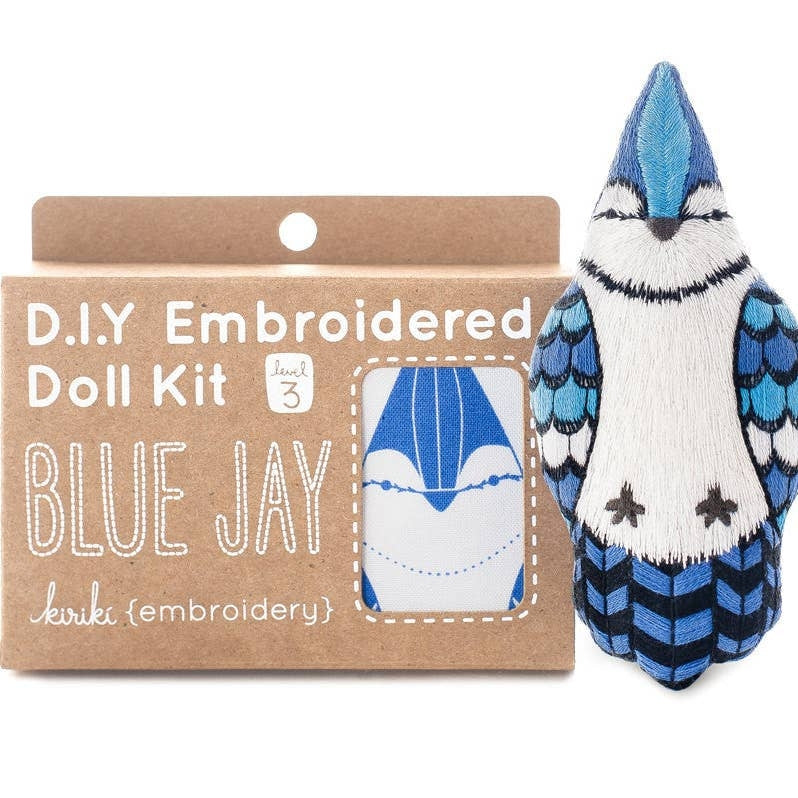 A DIY embroidered doll kit box next to a finished blue jay plush doll on a white background. The box is brown with white text. 