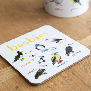 Coaster featuring illustrations of birds on a white background. Each bird is labelled, while the text 'Boobies' is printed on the top. The coaster sits on a wooden surface.