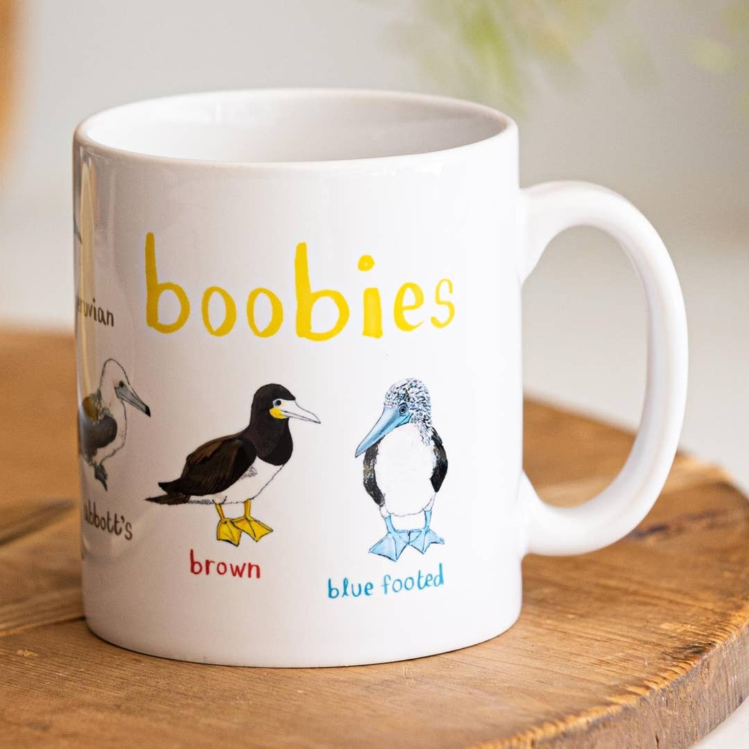 A white mug with bird illustrations. The text 'Boobies' is printed over the illustrations along with text labelling each image. The mug sits on a wooden surface.