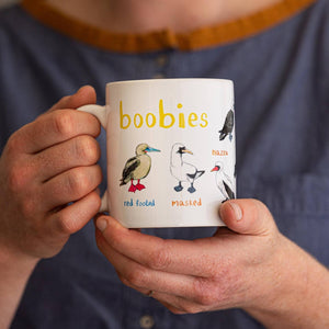 A person holding a white mug with bird illustrations. The text 'Boobies' is printed over the illustrations along with text labelling each image. 
