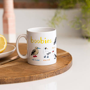 A white mug with bird illustrations. The text 'Boobies' is printed over the illustrations along with text labelling each image. The mug sits on a wooden surface.