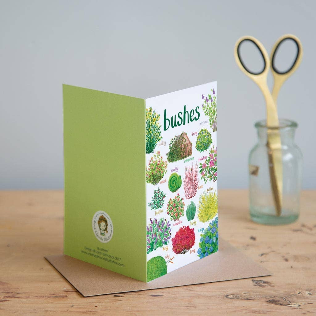 Greeting card featuring colourful illustrations of bushes on a white background. The heading 'Bushes' is printed at the top of the card while each of the illustrations is labelled with smaller text. The card has a green back and stands on a brown envelope on a wooden surface. 