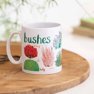 A white mug with colourful illustrations of different types of bushes. The text 'Bushes' is printed over the illustrations along with text labelling each image. The mug sits on a wooden surface. 