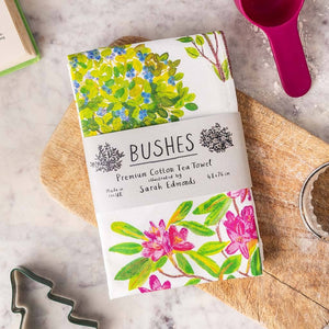 Image of a folded white cotton tea towel showing a colourful bush themed illustration. A paper label is wrapped around the centre of the towel with the text 'bushes', 'premium cotton tea towel', 'made in the UK' and 'illustrated by Sarah Edmonds' printed on it. The towel lies on a wooden board on a marble surface. 