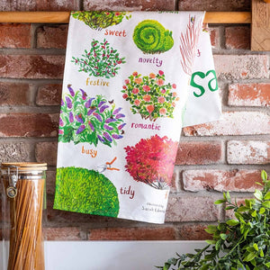 Image of a folded white cotton tea towel showing colourful illustrations of bushes with text printed underneath. The tea towel hangs on a wooden dowel against a red brick background. 