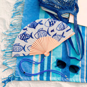 Image showing a handheld fan a blue fish design on a white background. The fan is attached to a blue lanyard to wear around your neck. It lies open on a blue striped beach towel on sand.