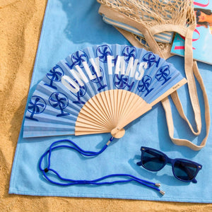 Open handheld fan with wooden ribs and a dark blue fan design and the text 'ONLY FANS' on a light blue background. The fan lies on a beach towel on top of sand, along with a bag and sunglasses.