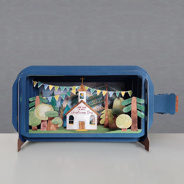 blue bottle pop up card. the intricate paper cuttings depict a forest scene with tree, flag banners, and a church with a text that reads "with love on your christening day" on its white panelling