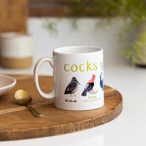 White mug with colourful bird themed illustrations. The text 'Cocks' is printed over the birds while different bird names are printed beside each illustration. The mug sits beside a spoon on a wooden board and white surface.