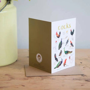 Greeting card featuring illustrations of cock birds on a white background. The heading 'cocks' is printed at the top of the card while each of the images are labelled with smaller text. The card stands on top of a brown envelope on a wooden surface.