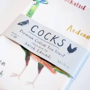 Close up of a folded cotton tea towel with a humorous bird-themed print featuring different types of cock birds, wrapped in a paper band that reads 'Cocks', 'Premium Cotton Tea Towel', and 'Illustrated by Sarah Edmonds'.