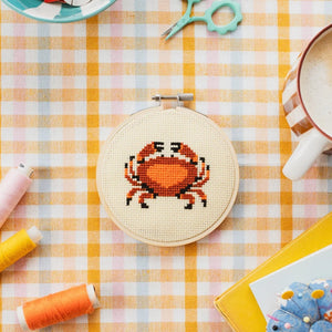 Cross-stitching hoop with an orange and red crab design on a checkered tablecloth with sewing supplies.