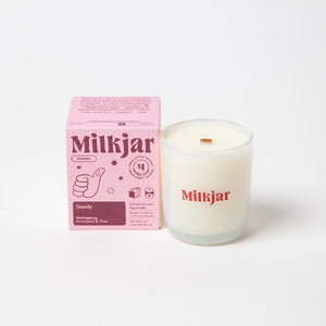 Canadian company Milkjar candle and its box packaging. The white candle is in a translucid glass with "Milkjar" written in pink. The other side of the candle has a cute thumbs up doodle in pink. The packaging is a small baby pink box with dark pink text and graphics. 