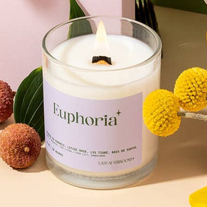 A candle in a milky-white glass jar filled with white wax and a lit wick. The text 'Euphoria' is printed on on a purple label on the candle, along with the company branding. The candles sits on a surface beside some acorns and flowers.