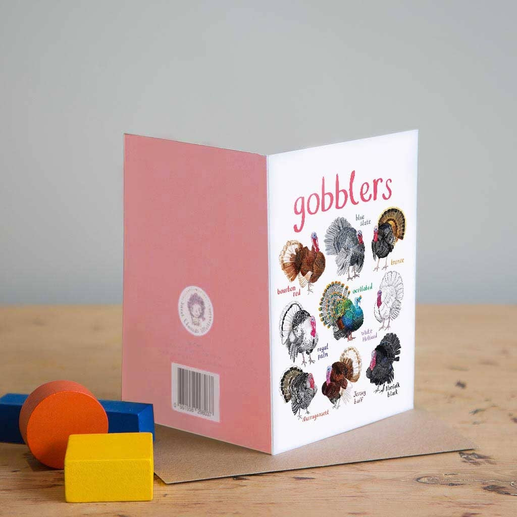 Greeting card with colourful illustrations of labelled turkeys on a white background. The text 'gobblers' is printed on the top of the card. The card sits on a wooden surface. 