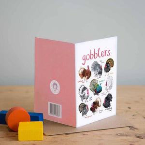 Greeting card with colourful illustrations of labelled turkeys on a white background. The text 'gobblers' is printed on the top of the card. The card sits on a wooden surface. 
