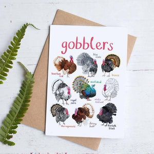 Greeting card with colourful illustrations of labelled turkeys on a white background. The text 'gobblers' is printed on the top of the card. The card sits with a brown envelope on a white background.