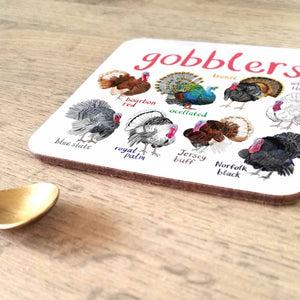 Coaster featuring illustrations of turkeys on a white background. Each turkey is labelled, while the text 'Gobblers' is printed on the top. The coaster sits on a wooden surface.