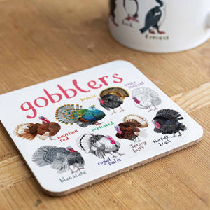 Coaster featuring illustrations of turkeys on a white background. Each turkey is labelled, while the text 'Gobblers' is printed on the top. The coaster sits on a wooden surface.