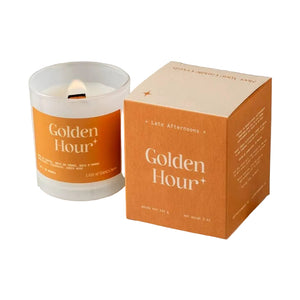 A candle in a milky-white glass jar filled with white wax and a lit wick. The candle sits next to an orange and cream packaging box on a white background. The text 'Golden Hour' is printed on both the candle and box, along with the company branding. 
