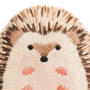 Close-up of a plush embroidered hedgehog face and upper body, featuring cream and brown threads for fur and spikes, black eyes and nose, and pink paw pads on a white background.