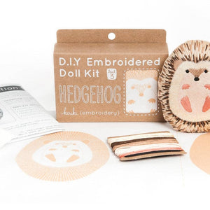 A DIY embroidered doll kit box next to a finished embroidered hedgehog doll plush on a white background. The box is brown with white text and sits behind a hedgehog embroidery pattern and some threads. 