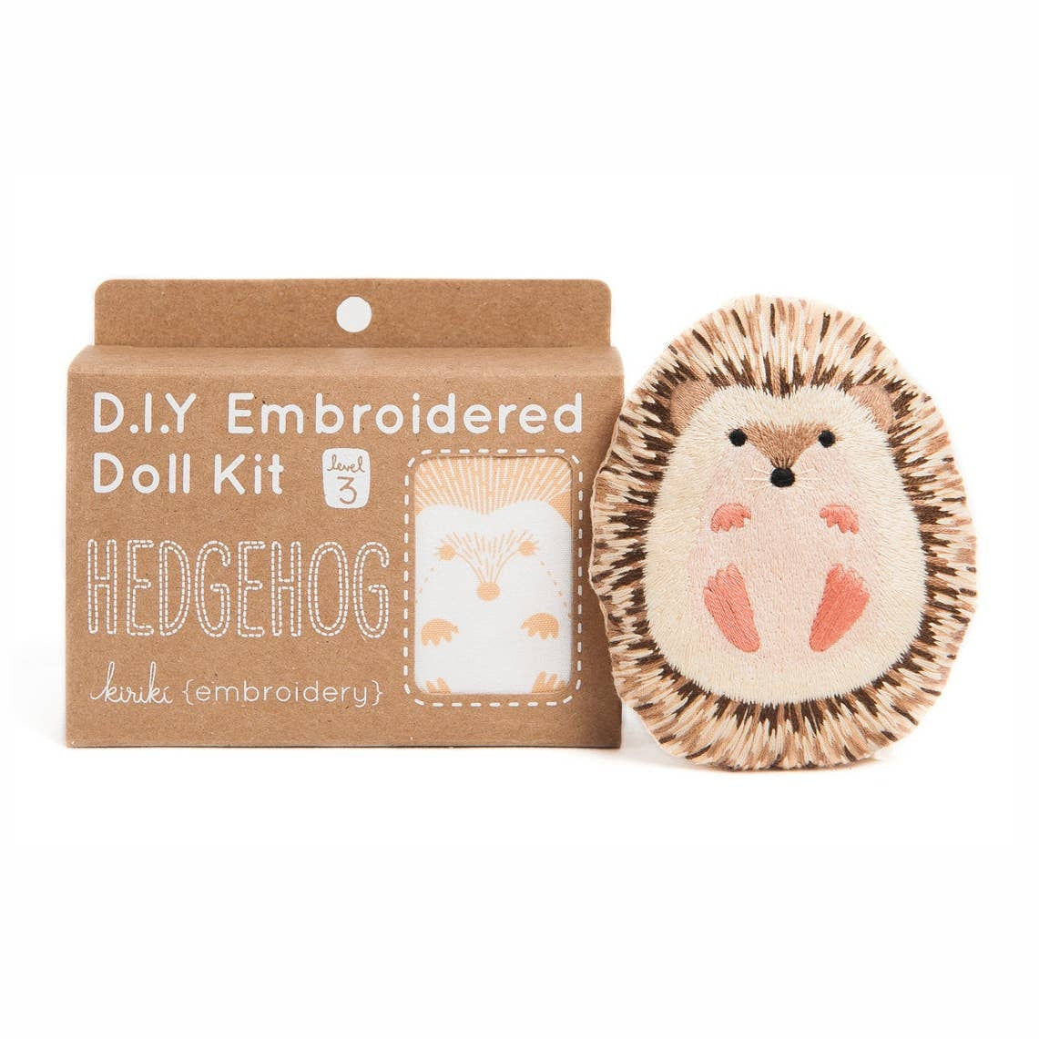 A DIY embroidered doll kit box next to a finished embroidered hedgehog doll plush on a white background. The box is brown with white text. 