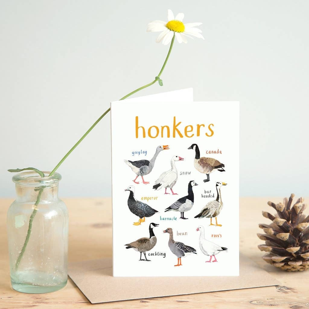Greeting card featuring illustrations of geese on a white background. The heading 'honkers' is printed at the top of the card while each of the images are labelled with smaller text. The card stands on top of a brown envelope on a wooden surface, beside a jar containing a flower, and a pine cone. 