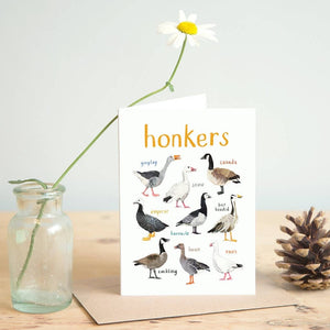 Greeting card featuring illustrations of geese on a white background. The heading 'honkers' is printed at the top of the card while each of the images are labelled with smaller text. The card stands on top of a brown envelope on a wooden surface, beside a jar containing a flower, and a pine cone. 