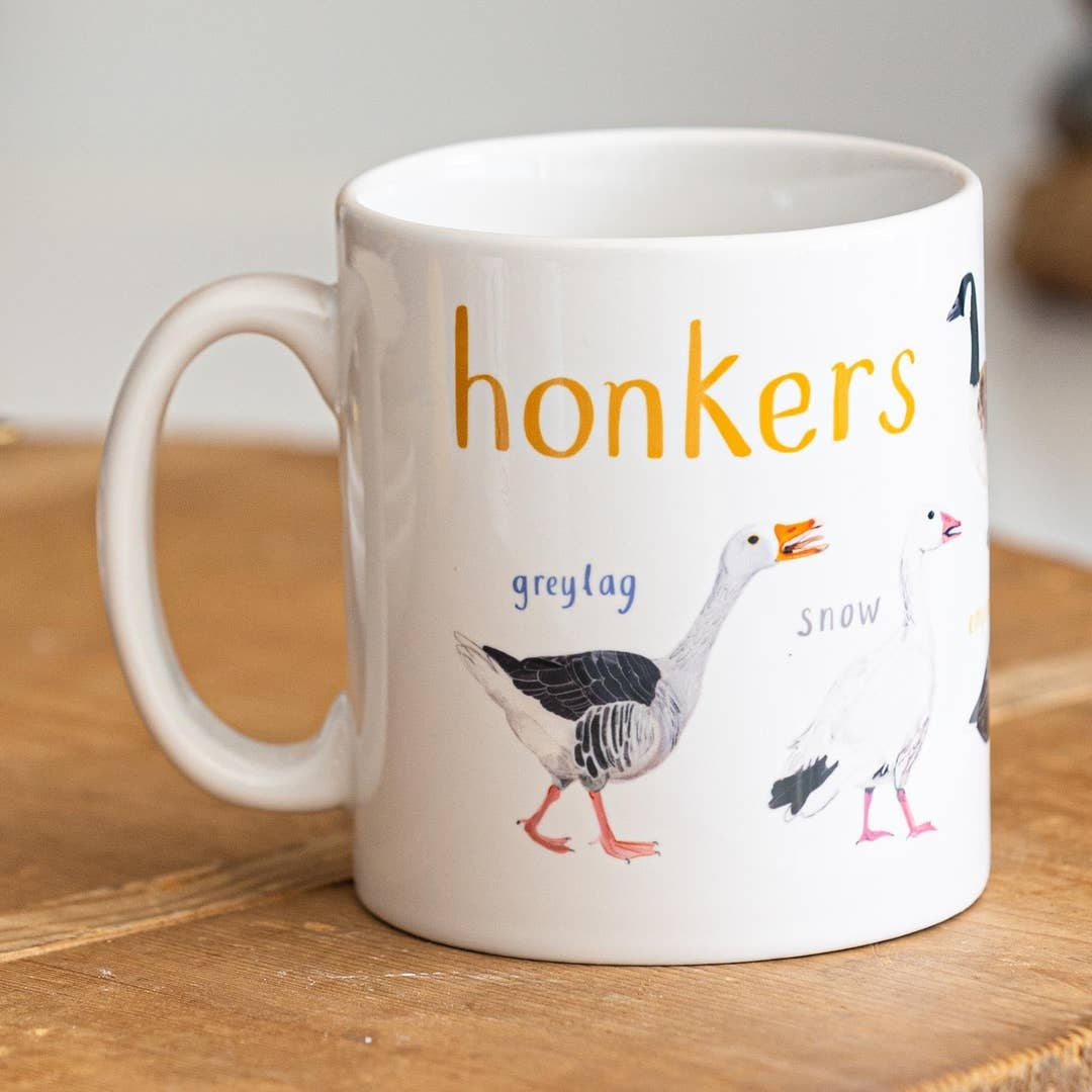 A white mug with geese illustrations. The text 'Honkers' is printed over the illustrations along with text labelling each image. The mug sits on a wooden surface.