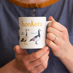 A person holding a white mug with geese illustrations. The text 'Honkers' is printed over the illustrations along with text labelling each image. 