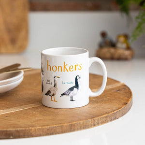 A white mug with geese illustrations. The text 'Honkers' is printed over the illustrations along with text labelling each image. The mug sits on a wooden surface.