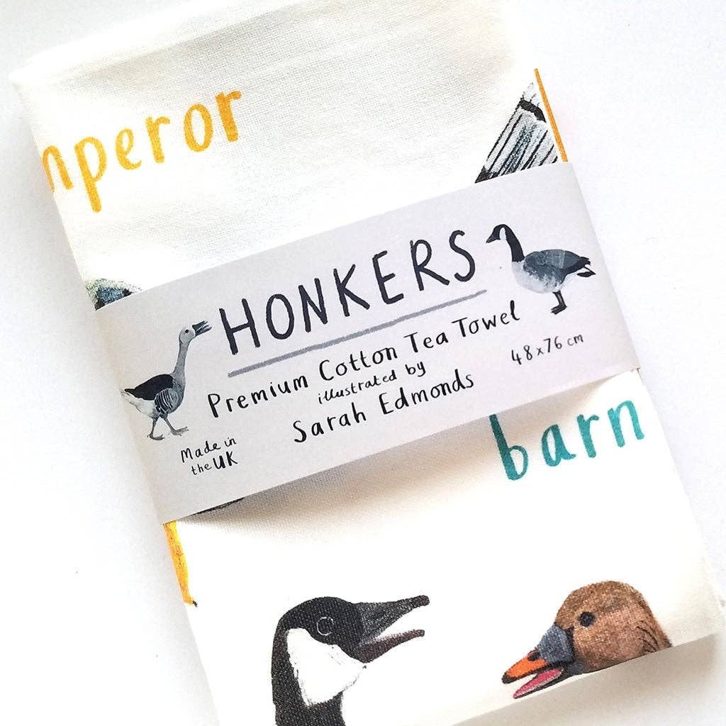 A white cotton tea towel showing colourful bird illustrations with text  printed underneath. The tea towel is folded and a paper label titled 'Honkers' is wrapped around the middle.