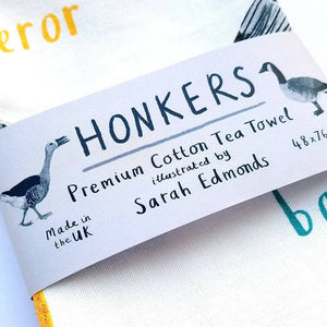 A close up of a paper label titled 'Honkers', wrapped around the middle of a tea towel. The label also shows two geese illustrations along with the text 'Premium Cotton Tea Towek', 'illustrated by Sarah Edmonds', and 'Made in the UK'.