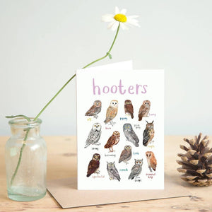 Greeting card featuring illustrations of owls on a white background. The heading 'hooters' is printed at the top of the card while each of the images is labelled with smaller text. The card stands beside a jar containing one flowers, and a pine cone on a wooden surface. 