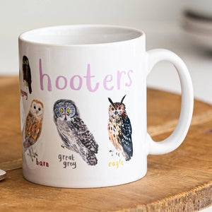 A white mug with owl illustrations. The text 'Hooters' is printed over the illustrations along with text labelling each image. The mug sits on a wooden surface.