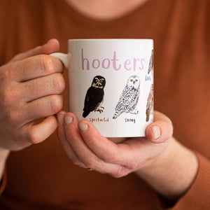 A person holding a white mug with owl illustrations. The text 'Hooters' is printed over the illustrations along with text labelling each image.