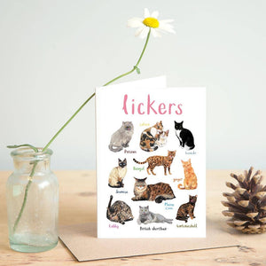 Greeting card featuring illustrations of cats on a white background. The heading 'lickers' is printed at the top of the card while each of the images are labelled with smaller text. The card stands on top of a brown envelope on a wooden surface, beside a jar containing a flower, and a pine cone. 