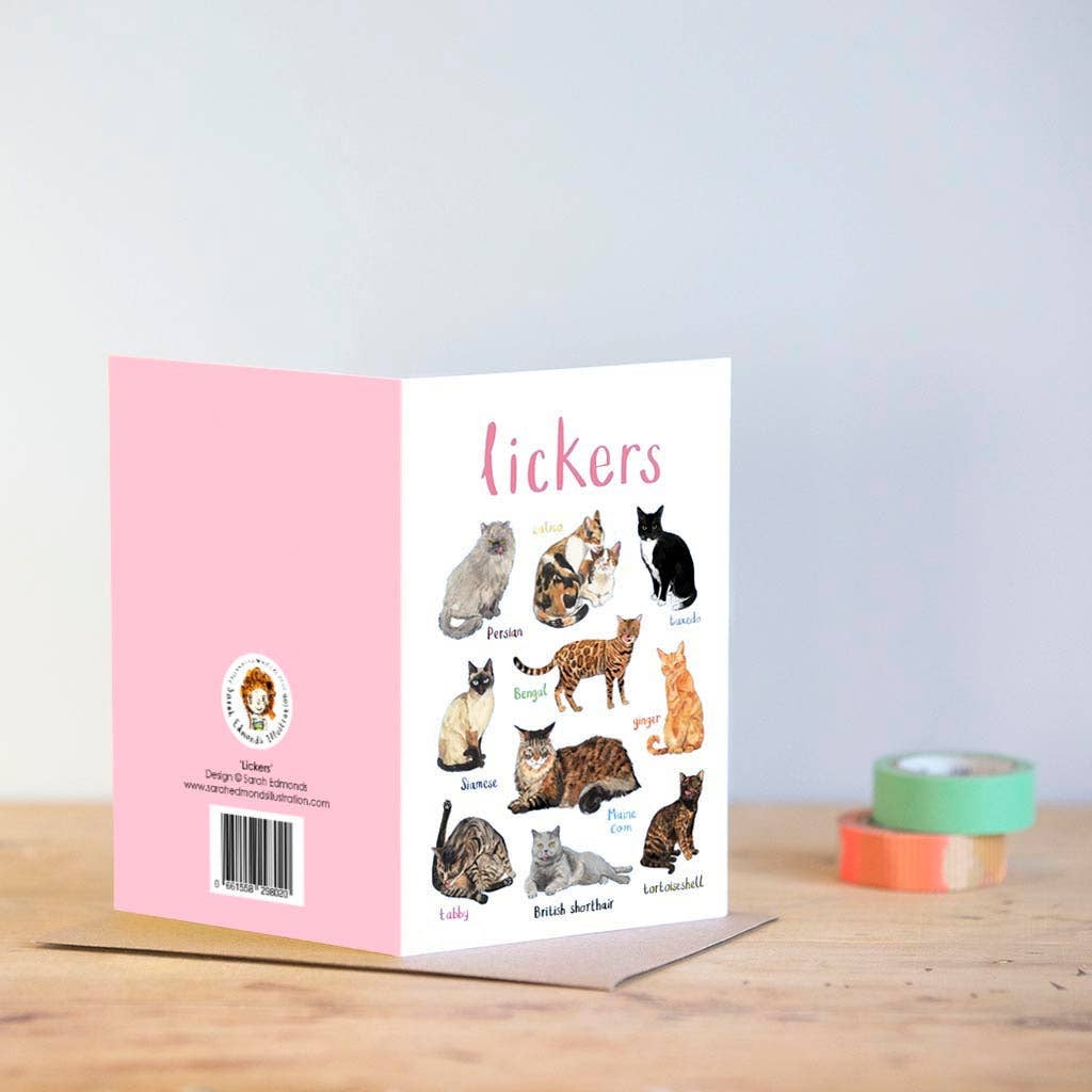 Greeting card featuring illustrations of cats on a white background. The heading 'lickers' is printed at the top of the card while each of the images are labelled with smaller text. The card stands on top of a brown envelope on a wooden surface, beside two rolls of tape.